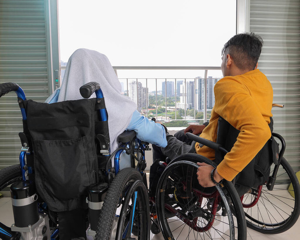 Юго-восточно-азиатский малайский мужчина женщина пара платка tudung среднего возраста инвалид на инвалидной коляске глядя сидя перед балконом окна смотреть на улицу держа крупным планом - Фото, изображение