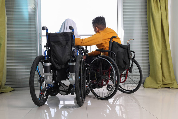 Güneydoğu Asya Malezyalı erkek çift başörtüsü Tudung tekerlekli sandalyede engelli balkon penceresinin önünde oturuyor. Dışarıya bak. - Fotoğraf, Görsel