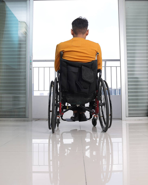 Südostasiatischer behinderter Mann mittleren Alters im Rollstuhl schaut vor Balkonfenster nach draußen - Foto, Bild
