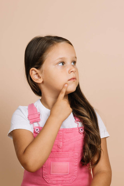 Portrait of child with shining bright eyes looking away, point finger on chin area of face with pensive look wearing bright pink jumpsuit and white t-shirt on beige background - Photo, image