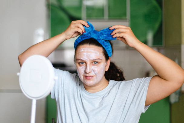 Young woman with face moisturizing mask nourishing skin adjusting blue bow on head. Talking care of yourself and skin. Youth extension - anti age. Home beauty makeup procedures - Φωτογραφία, εικόνα