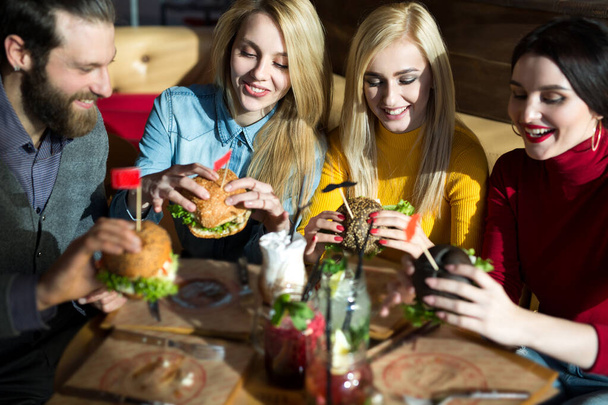 As pessoas jantam juntas numa mesa num café. Amigos felizes comem hambúrgueres e bebem coquetéis no restaurante
 - Foto, Imagem