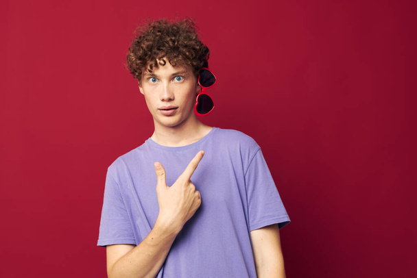 guy with red curly hair guy in a purple t-shirt with sunglasses posing red background - Fotó, kép