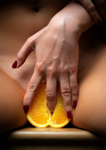 hermosa chica desnuda sexy sostiene una naranja en su abdomen inferior cerca de sus piernas. Sobre un fondo luminoso de luces. - Foto, Imagen