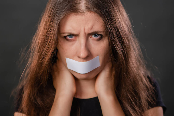 Close-up portrait of a young woman with duct tape sealed in her mouth, restriction of freedom of speech and censored and forbidden to speak and express her opinion, isolated on a dark background. - Valokuva, kuva