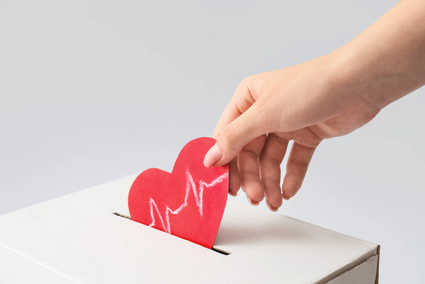 Femme mettant coeur de papier rouge avec dessin cardiogramme dans l'urne des dons sur fond clair, gros plan - Photo, image