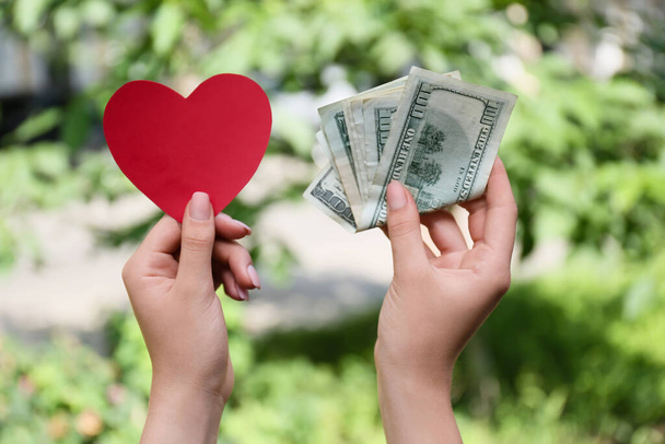 Γυναίκα που κρατάει κόκκινη χάρτινη καρδιά και χρήμα έξω, κοντινό πλάνο. Έννοια δωρεάς - Φωτογραφία, εικόνα