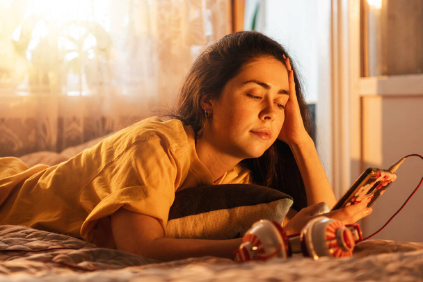 Egy csinos fiatal nő portréja fekszik az ágyon, zenét hallgat, fülhallgatót tesz a füléhez, belenéz az okostelefonjába. Napnyugta világítás az ablakból. - Fotó, kép