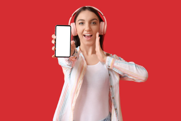 Здивована молода жінка в рожевих навушниках з мобільним телефоном на червоному тлі
 - Фото, зображення