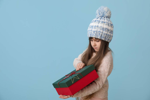 Cute little girl in winter clothes and with Christmas gift on color background - Photo, Image