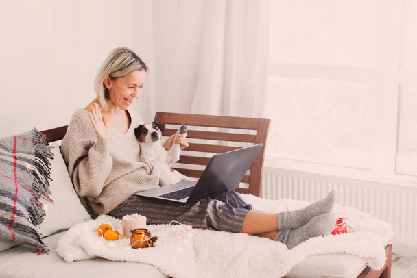 Happy middle-aged woman sit on sofa with dog at home video calling on laptop. Adult female browsing social media Video chat concept - Foto, afbeelding