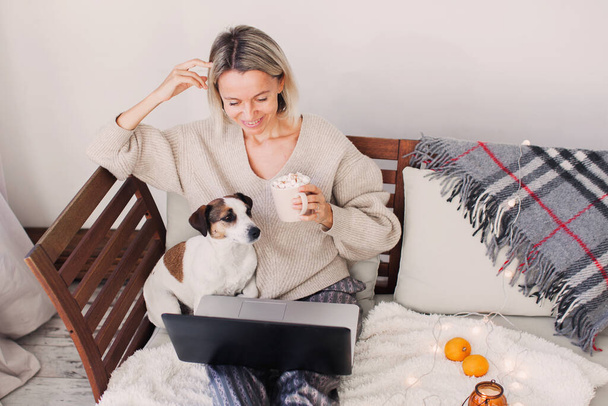 Charming middle-aged woman spends leisure time in networks on the computer, texting messages, watching movies. The adult lady works remotely with a laptop sitting on the sofa in cozy living room with dog - Zdjęcie, obraz