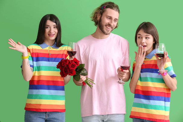 Man and two beautiful women in rainbow clothes, with glasses of wine and flowers on color background. Concept of polyamory and LGBT - Photo, Image