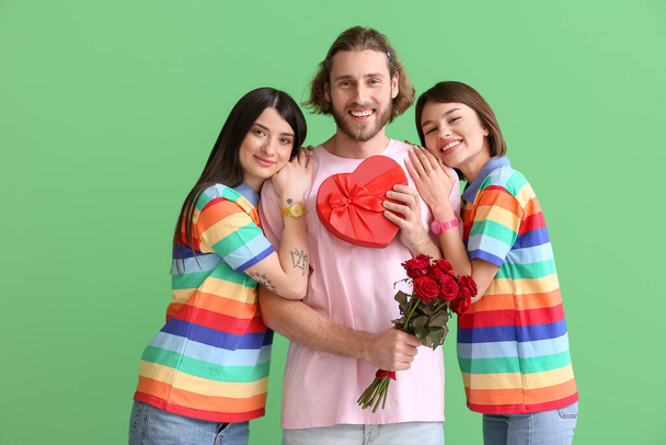 Man and two beautiful women in rainbow clothes, with gift and flowers on color background. Concept of polyamory and LGBT - Photo, Image