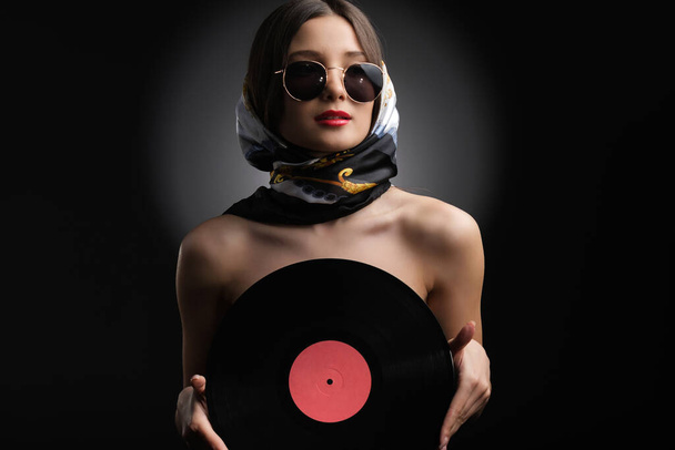 Fashionable pretty lady holding a vinyl record in her hands covering her chest wearing sunglasses and shawl on her head. - Foto, afbeelding
