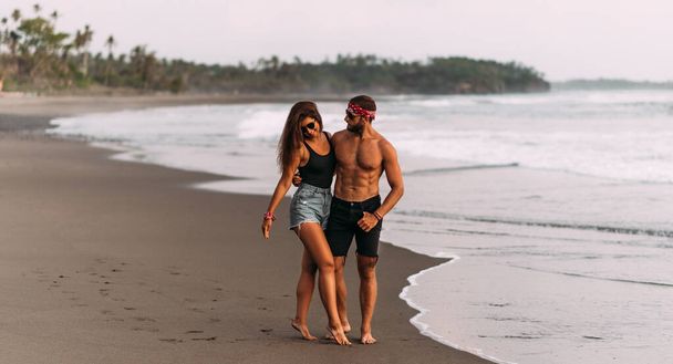 A tanned couple strolls along a sandy beach. A couple in love at sunset by the sea. A couple in love on vacation. Wedding trip. Romantic couple enjoying a walk on the beach at sunset. Copy space - Foto, Imagem