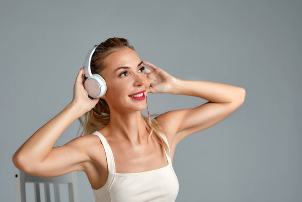 Banner of young happy girl in white swimsuit listening to music in white chair, wearing wireless headhones, isolated on gray background with copy space in studio - Foto, imagen