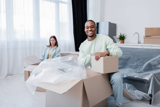 happy african american man unpacking box near blurred girlfriend  - Photo, Image