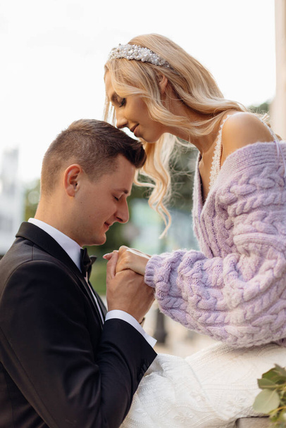 Side view of fair-haired bride with bandeau on her head wearing white wedding dress and lilac jacket and bridegroom in black suit with bow-tie standing close to her and taking each others hands - Foto, Imagen