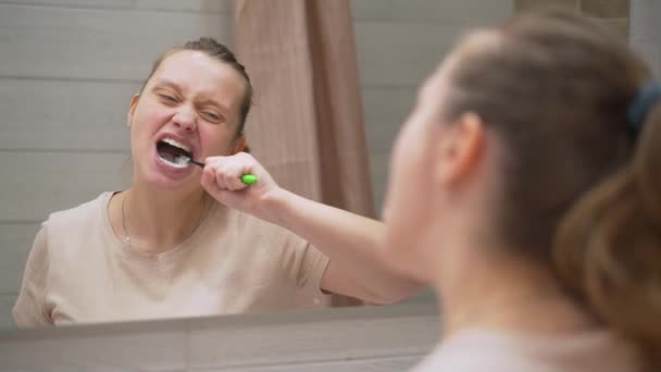 Woman in beige T-shirt actively brushes her teeth with green toothbrush in the bathroom at home. She cleans teeth from dirt and food for dental health. Healthy teeth, beautiful smile, daily routine - Filmati, video