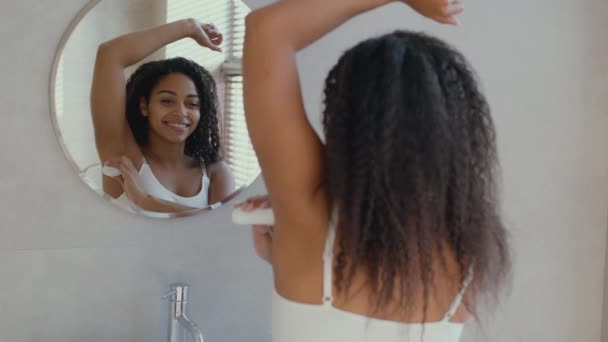 Hygiënische cosmetica. jonge afrikaanse amerikaanse vrouw aanbrengen stok deodorant op haar oksel, kijken naar spiegel op badkamer - Video