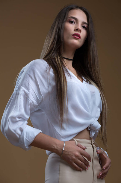 Portrait of a beautiful girl in a white shirt and leather leggings on a beige background - Фото, изображение