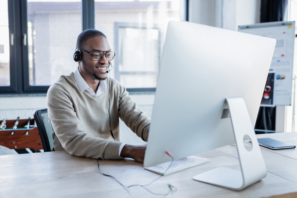 szczęśliwy afrykański operator amerykański w słuchawkach i okularach patrząc na monitor komputerowy w biurze  - Zdjęcie, obraz