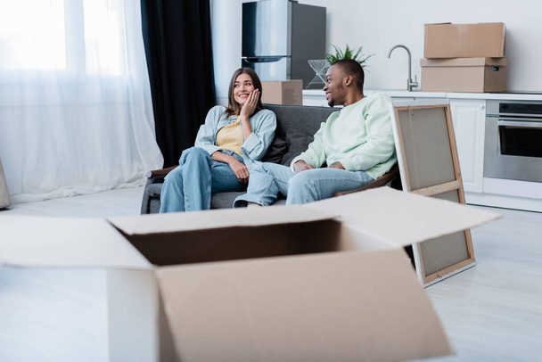 cheerful interracial couple sitting on sofa around carton boxes during relocation  - 写真・画像