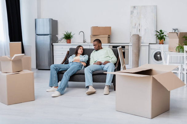 tired interracial couple sitting on couch around carton boxes during relocation  - Fotó, kép