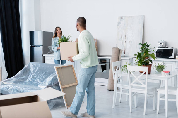 african american man holding carton box while looking at happy girlfriend with plant during relocation  - Foto, Imagen