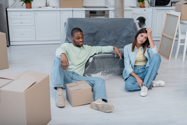displeased multiethnic couple sitting near boxes and sofa in plastic wrapper - Photo, Image