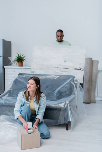 cheerful woman packing box with adjusting tape near african american man holding painting on blurred background - Foto, afbeelding