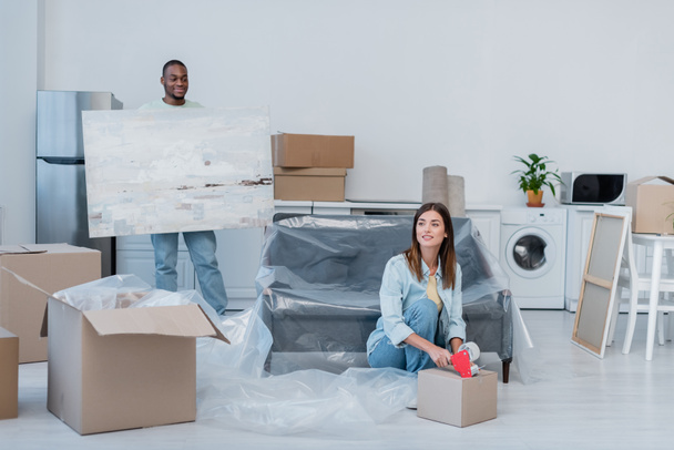 happy young woman packing box with adjusting tape near african american man holding painting in new home - Foto, immagini