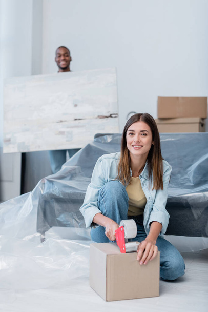 happy young woman packing box with adjusting tape near african american man holding painting on blurred background - Photo, Image