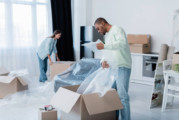 african american man holding plastic wrapper near box and blurred girlfriend during relocation  - Photo, Image