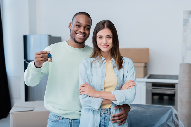 glücklicher afrikanisch-amerikanischer Mann hält Schlüssel in der Hand und umarmt zufriedene Freundin in neuer Wohnung  - Foto, Bild