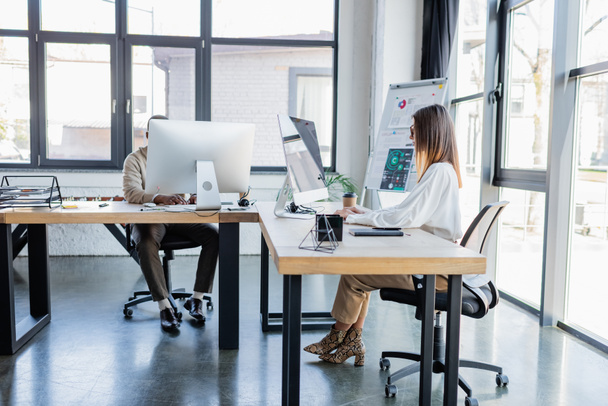 現代のオフィスでアフリカ系アメリカ人男性と仕事をしながらコンピュータモニターを見ている眼鏡のビジネス女性  - 写真・画像