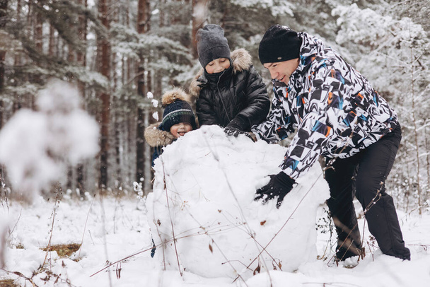 Happy father and sons rolling big snowball together for snowman in snowy winter forest. Man Joyful teenager and little boy playing and having fun on family walking. Wintertime activity outdoors. - Foto, Imagen