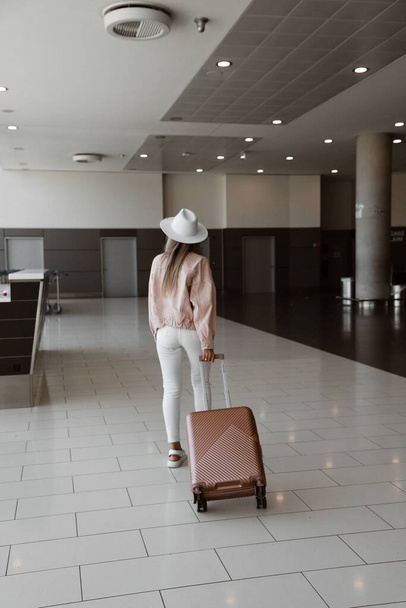 молодая девушка стоит в аэропорту перед отъездом, модель в белых брюках, розовая куртка и шляпа, розовый чемодан в руках, женщина идет назад - Фото, изображение