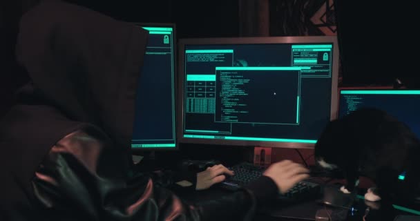Hacker σε μια κουκούλα κάθεται μπροστά από οθόνες υπολογιστών και αμυχές βάσεις δεδομένων - Πλάνα, βίντεο
