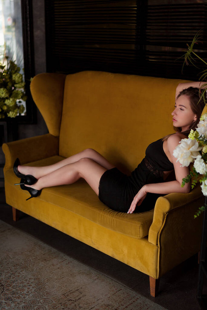 sexy attraktive Frau in einem schwarzen Kleid in schwarzen Schuhen sitzt auf einem gelben Sofa, vertikales Foto - Foto, Bild