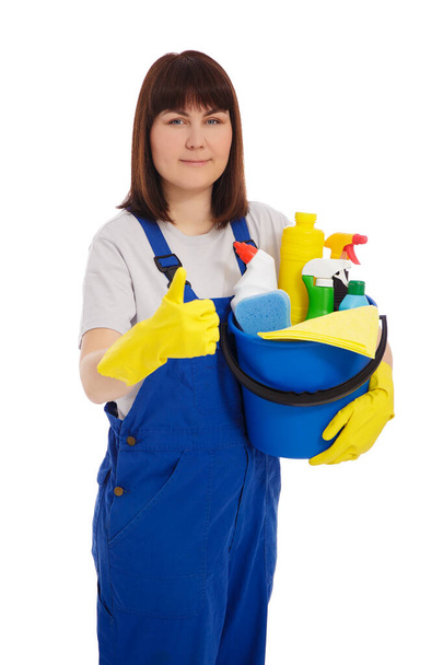 Hausarbeit und Reinigungskonzept - Porträt einer jungen Putzfrau in blauer Uniform und gelben Handschuhen, die einen Eimer mit Putzutensilien hält und vereinzelt die Daumen nach oben auf weißem Hintergrund hebt - Foto, Bild