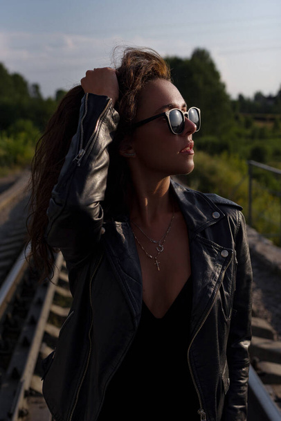  молодая сексуальная брюнетка в черном комбинезоне, кожаной куртке и солнечных очках - Фото, изображение