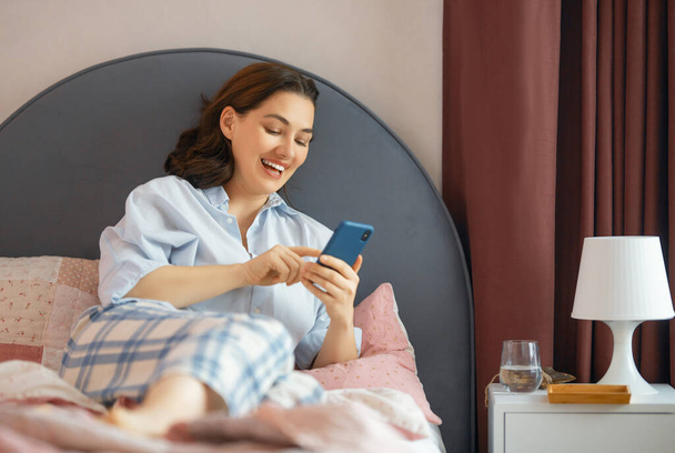 Happy casual beautiful woman is using a phone sitting on a bed at home. - Fotografie, Obrázek