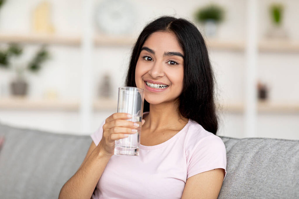 Sorrindo jovem indiana segurando vidro de água limpa fresca enquanto sentado no sofá na sala de estar. Mantenha o conceito hidratado - Foto, Imagem