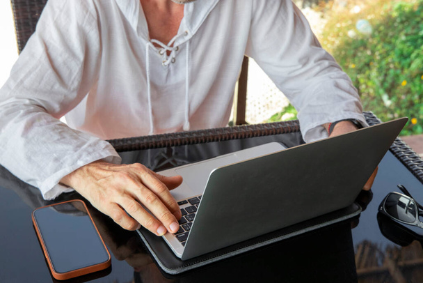 Férfi szabadúszó dolgozik egy laptop, gépelés a billentyűzeten, miközben ül az asztalnál a beach cafe. A szabadban dolgozom. Szabadúszó koncepció - Fotó, kép