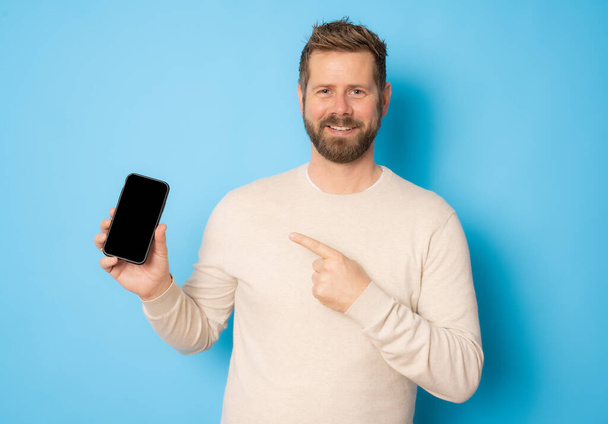Portrait of a cheerful young man wearing casual clothing isolated over blue background, showing blank screen mobile phone - Fotoğraf, Görsel