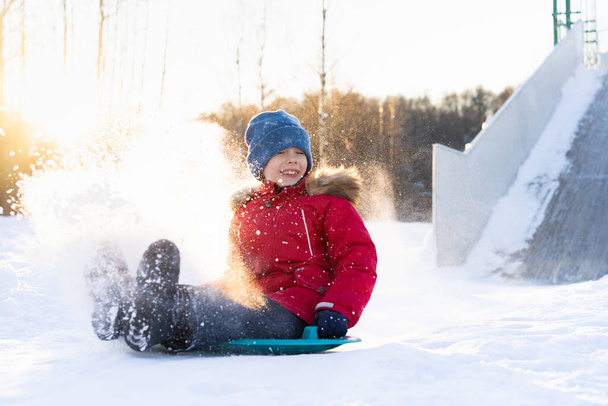 malé dítě v červené bundě sjíždí po skluzavce na ledu, veselé dítě, emoce ze zimní zábavy. - Fotografie, Obrázek