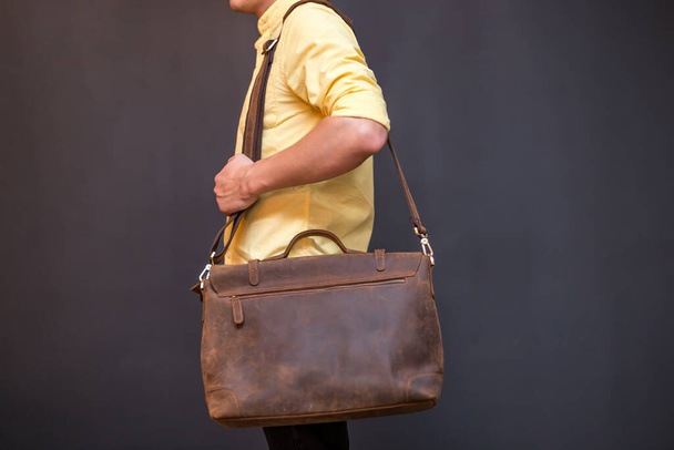 Man carries brown leather messenger bag in the hand on gray background. Unisex bag for sale. - Foto, imagen