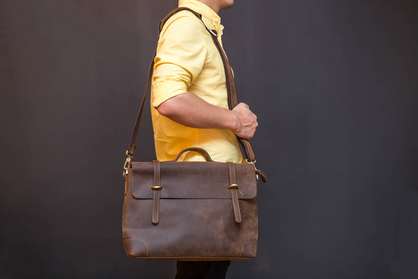 Man carries brown leather messenger bag in the hand on gray background. Unisex bag for sale. - Fotografie, Obrázek